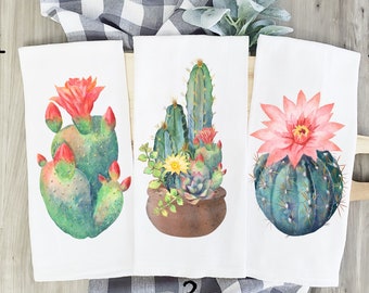 Puede incluir: Tres toallas de cocina con diferentes diseños de cactus de acuarela. La primera toalla presenta un cactus de chumbera con flores rosas. La segunda toalla presenta un cactus alto con flores verdes y amarillas. La tercera toalla presenta un cactus redondo con una flor rosa.