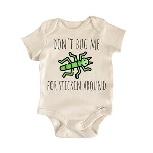 Op de afbeelding: Crèmekleurige babybody met de tekst "DON'T BUG ME FOR STICKIN AROUND" in zwart. Een groen en zwart cartoon insect is gecentreerd boven de tekst. De body is gemaakt van zacht materiaal en heeft korte mouwen.