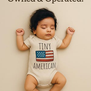 A sleeping baby wearing a white onesie with the text "Tiny American" printed on it.