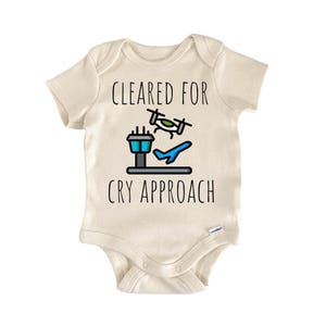 May include: Cream-colored baby onesie with the text "CLEARED FOR CRY APPROACH" in black. The design includes a blue airplane, a green airplane, and a blue and black control tower graphic. A cute and funny baby outfit.