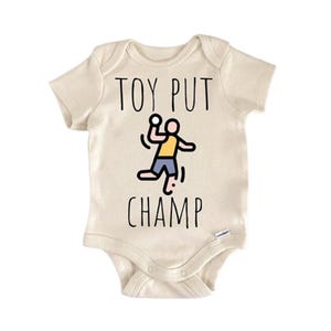 Könnte beinhalten: Cremefarbener Baby-Body mit dem schwarzen Text "TOY PUT CHAMP". In der Mitte befindet sich eine Cartoonfigur, die einen Ball wirft. Die Figur trägt ein gelbes Hemd und blaue Shorts.