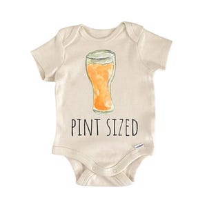 Puede incluir: Un body de bebé color crema con una ilustración de acuarela de una pinta de cerveza. El texto "PINT SIZED" está impreso en negro debajo del gráfico de la cerveza. El body tiene mangas cortas y cierres de broche.