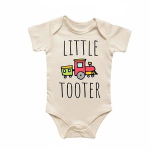 Könnte beinhalten: Ein cremefarbener Baby-Body mit dem Schriftzug "LITTLE TOOTER" in schwarzen Buchstaben. Eine farbenfrohe Zugillustration in Rot, Gelb, Grün und Blau befindet sich unter dem Wort "LITTLE".
