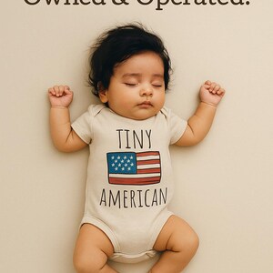 A sleeping baby wearing a white onesie with the text "Tiny American" printed on it.