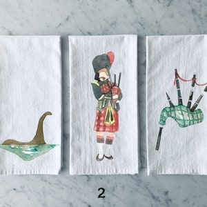 Può includere: Tre asciugamani da cucina bianchi con diversi disegni. Il primo asciugamano presenta un'illustrazione ad acquerello marrone e verde del mostro di Loch Ness. Il secondo asciugamano presenta un'illustrazione ad acquerello di una persona che indossa un kilt e suona la cornamusa. Il terzo asciugamano presenta un'illustrazione ad acquerello di un set di cornamuse.