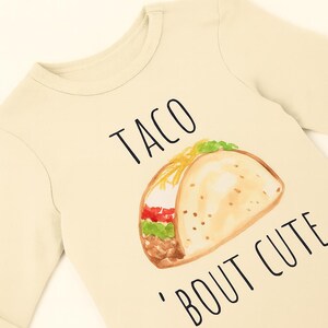 A beige long-sleeved t-shirt with a printed design of a taco and the text "TACO 'BOUT CUTE" printed on it.