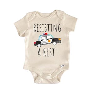 Può includere: Body da bambino color crema con la scritta "RESISTING A REST" e un'illustrazione ad acquerello di un'auto della polizia. L'auto della polizia è blu, bianca, nera e gialla.
