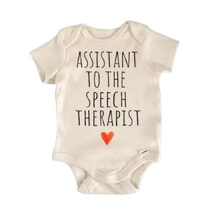 May include: A cream-colored baby onesie with the text "ASSISTANT TO THE SPEECH THERAPIST" in black, with a small red heart below the text. The onesie has short sleeves and snaps at the bottom.