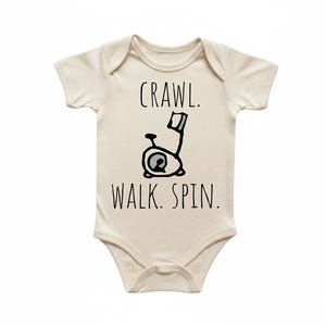 May include: Cream-coloured baby onesie with the text "Crawl. Walk. Spin." above and below a black and white illustration of a stationary exercise bike. The onesie has short sleeves and a round neckline.