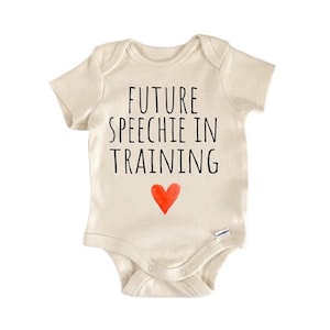 May include: Cream-colored baby onesie with the text "FUTURE SPEECHIE IN TRAINING" in black, and a red heart. The onesie has short sleeves and snaps at the bottom. A cute and playful design for a baby.