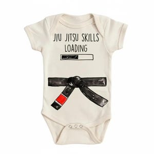 Op de afbeelding: Crème babyromper met de tekst "JIU JITSU SKILLS LOADING" boven een laadbalk. Een zwarte band met een rode streep is eronder afgebeeld. De romper heeft korte mouwen en drie drukkers aan de onderkant.
