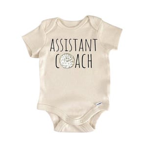 Può includere: Body da bambino beige chiaro con la scritta "ASSISTANT COACH" in nero. Una grafica di pallina da golf è centrata sotto il testo. Il body ha maniche corte e chiusura con bottoni automatici.