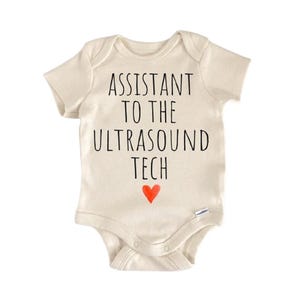 Può includere: Body da bambino color crema con la scritta nera "ASSISTANT TO THE ULTRASOUND TECH" e un piccolo cuore rosso. Il body ha maniche corte e bottoni automatici sul fondo.
