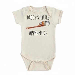 Puede incluir: Body de bebé color crema con el texto "DADDY'S LITTLE APPRENTICE" en negro. Una ilustración de acuarela de una llave inglesa está impresa encima de la palabra "APPRENTICE". El body tiene mangas cortas y corchetes en la parte inferior.