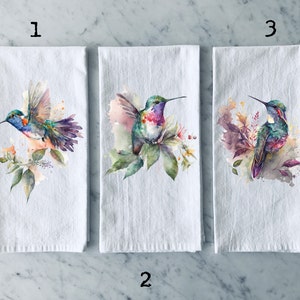 May include: Three white tea towels with watercolor hummingbird designs. Each towel features a different hummingbird with colorful feathers and floral accents.