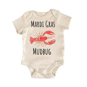 Può includere: Un body per neonati a maniche corte color crema con un'illustrazione di un gambero rosso decorato con perline colorate. Il testo "MARDI GRAS" è stampato in nero sopra e la parola "MUDBUG" è stampata in nero sotto.
