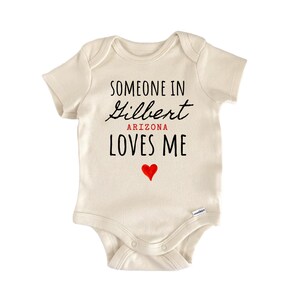 May include: An off-white short-sleeve baby bodysuit featuring the black text SOMEONE IN, a script font for Gilbert, red text for ARIZONA, black text for LOVES ME, and a small red heart graphic centered below the text on the front of the garment.