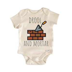 Puede incluir: Body de bebé color crema con las palabras "DROOL AND MORTAR" en negro. El diseño incluye un muro de ladrillos y una llana. El body está hecho de material suave y tiene mangas cortas.