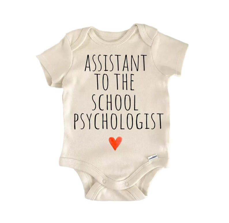 A beige baby onesie with the text "ASSISTANT TO THE SCHOOL PSYCHOLOGIST" printed on it, along with a small red heart symbol.
