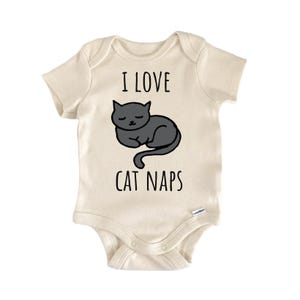 May include: A cream-colored short-sleeve bodysuit featuring a graphic of a sleeping dark gray cat in the center. The text "I LOVE CAT NAPS" is printed in black lettering above and below the cat illustration. The garment has a round neckline.