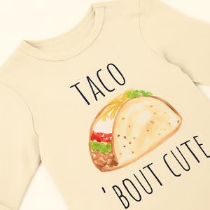 A beige long-sleeved t-shirt with a printed design of a taco and the text "TACO 'BOUT CUTE" printed on it.