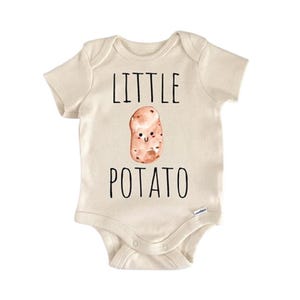 Puede incluir: Body de bebé color crema con las palabras "LITTLE POTATO" en letras negras. Una ilustración de acuarela de una patata sonriente está centrada entre las palabras. El body tiene mangas cortas y cierres de presión.