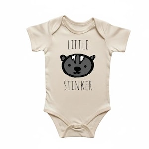 May include: Cream-colored baby bodysuit with a cartoon skunk face and the words "LITTLE STINKER" in black. The skunk has a black outline, gray face, and white stripe.