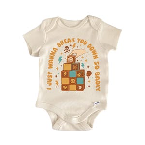 May include: Cream-colored baby bodysuit with a graphic design. The design features stacked blocks with various symbols, including a skull, lightning bolts, and a broken heart. The text reads "I JUST WANNA BREAK YOU DOWN SO BADLY."