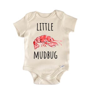 May include: A cream-colored short-sleeve bodysuit featuring a watercolor illustration of a red crawfish in the center. The text "LITTLE" is printed in black above the crawfish, and the text "MUDBUG" is printed in black below the illustration.
