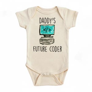 Puede incluir: Body de bebé color crema con el texto "DADDY'S FUTURE CODER" encima y debajo de un gráfico de ordenador estilo acuarela con una pantalla turquesa y símbolos de codificación.