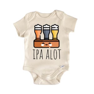 Puede incluir: Body de bebé color crema con el texto negro "IPA ALOT". El gráfico presenta tres vasos de cerveza en naranja, gris y amarillo, con espuma blanca, sobre una bandeja marrón con una cara sonriente. Un lindo atuendo para bebé.
