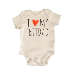 Op de afbeelding: Een crèmekleurige baby-onesie met korte mouwen. De tekst "I ♥ MY EBITDAD" is op de voorkant gedrukt, met een rood hart dat het woord "love" vervangt. De onesie heeft drukknoopsluitingen aan de onderkant.