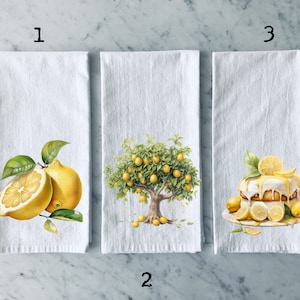 May include: Three white kitchen towels with lemon-themed designs. The first towel features a single lemon cut in half, the second towel features a lemon tree with yellow lemons, and the third towel features a lemon cake with white frosting and yellow lemon slices.