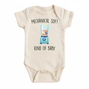 May include: Cream-colored baby bodysuit with the text "MECHANICAL SOFT KIND OF BABY". The graphic shows a blue blender filled with fruit. A cute and funny baby shower gift.