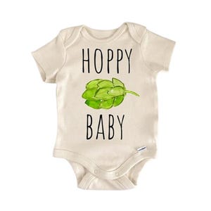 Puede incluir: Body de bebé color crema con las palabras "HOPPY BABY" en letras negras. Una ilustración de lúpulo verde está centrada entre las palabras. El body tiene mangas cortas y corchetes en la parte inferior.