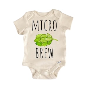 Puede incluir: Body de bebé color crema con las palabras "MICRO BREW" en letras negras. Una ilustración de acuarela verde de un cono de lúpulo está centrada entre las palabras. El body tiene mangas cortas y corchetes en la parte inferior.