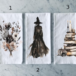 Könnte beinhalten: Drei Küchentücher mit verschiedenen Halloween-Motiven. Das erste Handtuch zeigt einen Aquarell-Blumenstrauß in Braun-, Schwarz- und Weißtönen. Das zweite Handtuch zeigt eine Aquarell-Illustration einer Hexe in einem schwarzen Kleid und Hut. Das dritte Handtuch zeigt eine Aquarell-Illustration eines Bücherstapels mit einer Kerze und einem Kürbis.