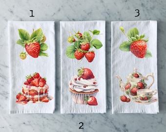 Puede incluir: Tres toallas de cocina blancas con diseños de fresas. La primera toalla presenta una sola fresa con hojas verdes. La segunda toalla presenta un cupcake con glaseado blanco y fresas rojas. La tercera toalla presenta una tetera con un diseño de fresa y una taza y un platillo.