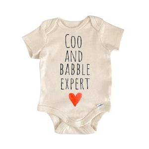 May include: Cream-colored baby onesie with the text "COO AND BABBLE EXPERT" in black, and a red heart at the bottom. The onesie has short sleeves and snaps at the bottom for easy changes. A cute and playful baby garment.