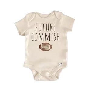 Puede incluir: Body de bebé color crema con el texto negro "FUTURE COMMISH" sobre un gráfico de fútbol americano marrón. El body tiene mangas cortas y cierres de broche.