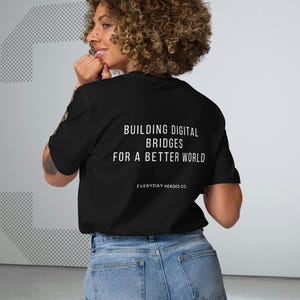 Peut inclure: T-shirt noir avec texte blanc : « BUILDING DIGITAL BRIDGES FOR A BETTER WORLD » et « EVERYDAY HEROES CO. » La personne porte un jean bleu clair. Le fond est blanc avec une grande forme grise.