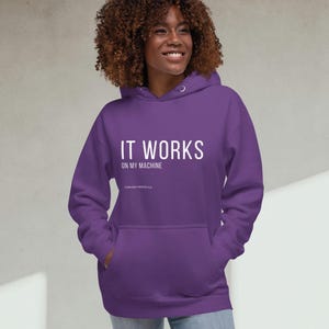 Può includere: Una felpa con cappuccio viola con la scritta bianca "IT WORKS ON MY MACHINE". La felpa ha un cappuccio con coulisse e una tasca frontale. La modella indossa jeans blu.