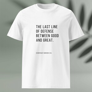 May include: A white t-shirt featuring the black text "THE LAST LINE OF DEFENSE BETWEEN GOOD AND GREAT." with "EVERYDAY HEROES CO." printed below. The shirt has a crew neck and short sleeves, a classic design.