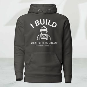 以下が含まれることがあります： フロントポケットとドローストリングフードが付いた濃いグレーのパーカー。「I BUILD WHAT OTHERS DREAM」という白い文字と、建設作業員のグラフィックが描かれています。ブランド名「EVERYDAY HEROES CO.」が下にあります。