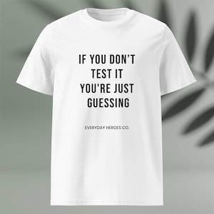 Op de afbeelding: Wit t-shirt met de zwarte tekst "IF YOU DON'T TEST IT YOU'RE JUST GUESSING" en de merknaam "EVERYDAY HEROES CO.". Het shirt heeft een ronde hals en korte mouwen.