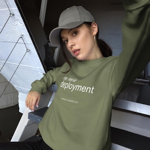 May include: An olive green sweatshirt with the words "one more deployment" in white. The sweatshirt also features the text "EVERYDAY HEROES CO." below. The person in the image is wearing a grey baseball cap.