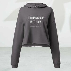 May include: A charcoal gray cropped hoodie with long sleeves. The hoodie features the text "TURNING CHAOS INTO FLOW" and "EVERYDAY HEROES CO." in white. The bottom hem is raw-edged.