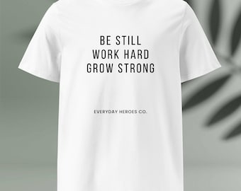 May include: A white t-shirt featuring the black text "BE STILL WORK HARD GROW STRONG" in a bold font, centrally placed. Beneath this, in a smaller font, is "EVERYDAY HEROES CO." The shirt has a round neck and short sleeves.