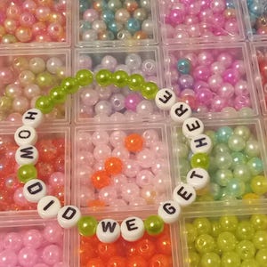 May include: A bracelet spelling "HOW DID WE GET HERE" with white letter beads and green beads. The bracelet is set against a backdrop of various coloured beads in a segmented container.