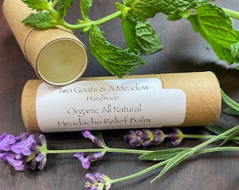 May include: A brown cardboard tube with a white label that reads "Two Goats & A Meadow Handmade Organic All Natural Headache Relief Balm". The tube is surrounded by green mint leaves and purple lavender flowers.